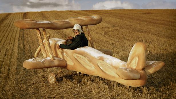 Ein Mann in Fliegerhaube sitzt in einem Flugzeug, das aus Brot geformt ist, auf einem Feld.