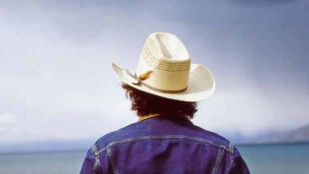 Ein Mann mit Cowboyhut und Jeansjacke blickt auf einen See.
