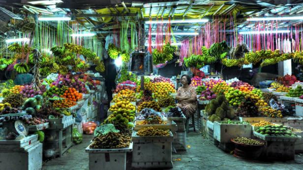 Eine Frau sitzt inmitten eines farbenfrohen Obstmarktes mit einer großen Auswahl an Früchten.