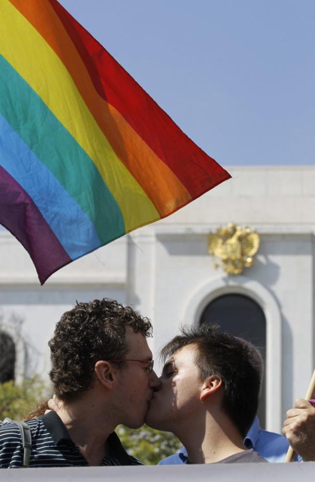 Ein schwules Paar küsst sich vor einer Regenbogenflagge und einem Gebäude mit einem goldenen Wappen.