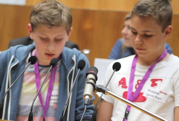 Zwei Jugendliche stehen an einem Rednerpult mit Mikrofonen und tragen Namensschilder „Pressesprecher“.