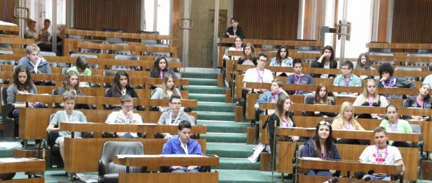Eine Gruppe junger Leute sitzt in einem großen Saal mit Holzbänken.