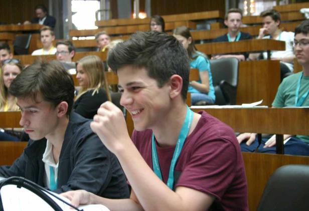 Ein lächelnder junger Mann sitzt in einem Hörsaal mit anderen Studenten.