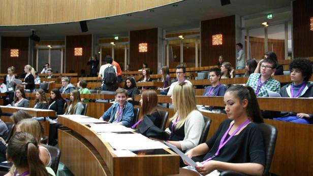Eine Gruppe junger Menschen sitzt in einem großen Saal mit Holzbänken.