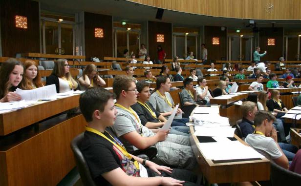 Jugendliche sitzen in einem großen Saal mit Holzbänken und lesen Papiere.