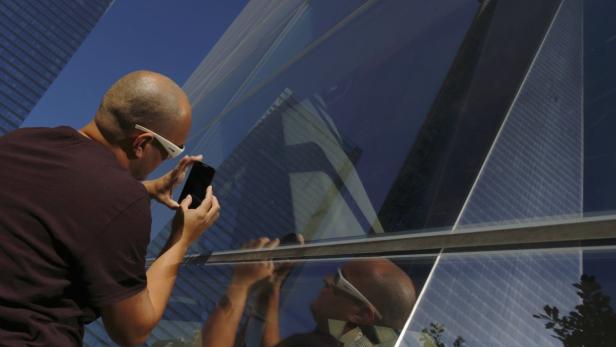 Ein Mann mit weißer Brille fotografiert ein Gebäude, das sich in einer Glasfassade spiegelt.