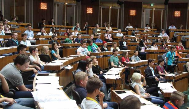 Zahlreiche junge Menschen sitzen in einem großen Saal mit Holzbänken.