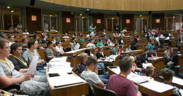 Viele junge Menschen sitzen in einem großen Hörsaal mit hölzernen Tischen.