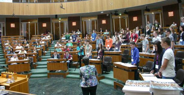 Eine Gruppe junger Menschen steht in einem großen Saal mit hölzernen Bänken und Pulten.