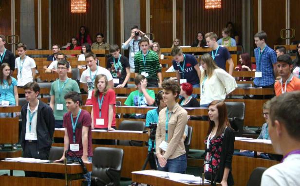 Eine Gruppe Jugendlicher sitzt oder steht in einem großen Saal mit Holztäfelung.