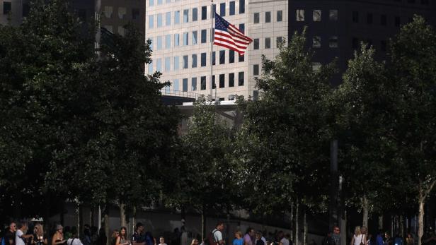 Eine amerikanische Flagge weht vor einem Bürogebäude, gesehen durch Baumreihen und Menschen.