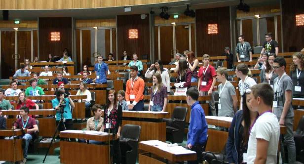 Eine Gruppe junger Leute sitzt und steht in einem großen Saal mit Holzbänken.