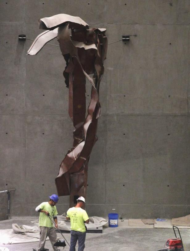 Zwei Bauarbeiter reinigen den Bereich um eine Skulptur aus Stahlträgern vor einer Betonwand.