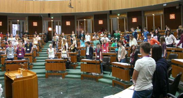 Eine Gruppe junger Menschen steht in einem großen, holzgetäfelten Saal an ihren Plätzen.