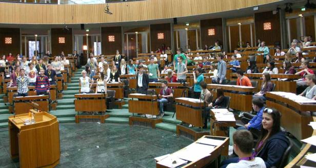 Viele junge Menschen sitzen in einem großen, holzvertäfelten Saal mit Pulten.