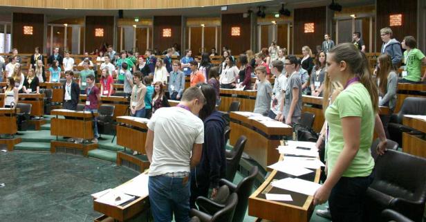 Eine Gruppe junger Leute steht in einem großen Saal mit Holztischen und Stühlen.