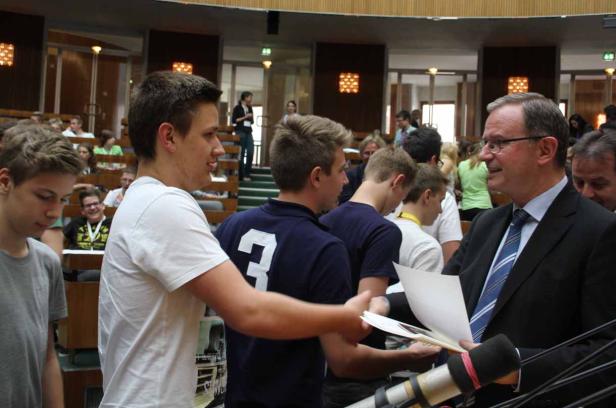 Ein Mann im Anzug überreicht Dokumente an eine Gruppe junger Leute in einem Hörsaal.