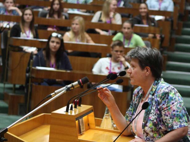 Eine Frau spricht vor einem Publikum an einem Rednerpult mit mehreren Mikrofonen.