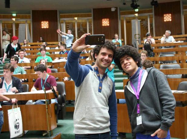 Zwei junge Männer machen ein Selfie in einem großen Saal mit vielen Sitzplätzen.