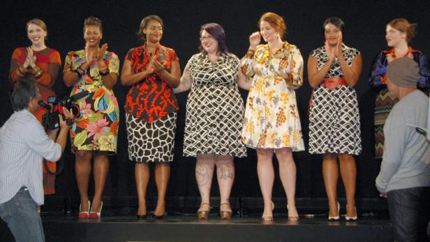 Sieben Frauen in farbenfrohen Kleidern stehen auf einer Bühne und applaudieren.