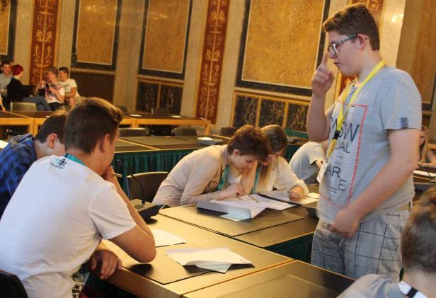 Junge Leute sitzen an Tischen und arbeiten an Dokumenten in einem großen Raum.