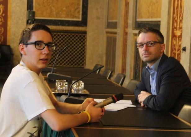 Zwei Männer sitzen an einem Tisch mit Mikrofonen in einem formellen Raum.