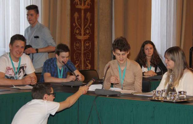 Eine Gruppe junger Leute sitzt an einem Tisch mit Mikrofonen und nimmt an einer Konferenz teil.