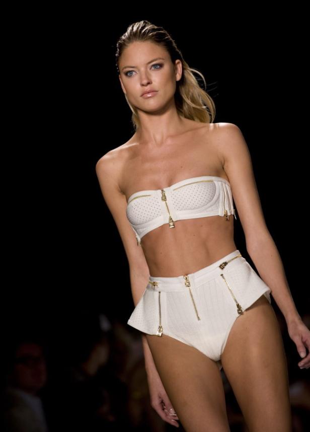 Eine Frau präsentiert ein weißes zweiteiliges Outfit mit goldenen Reißverschlüssen auf einem Laufsteg.