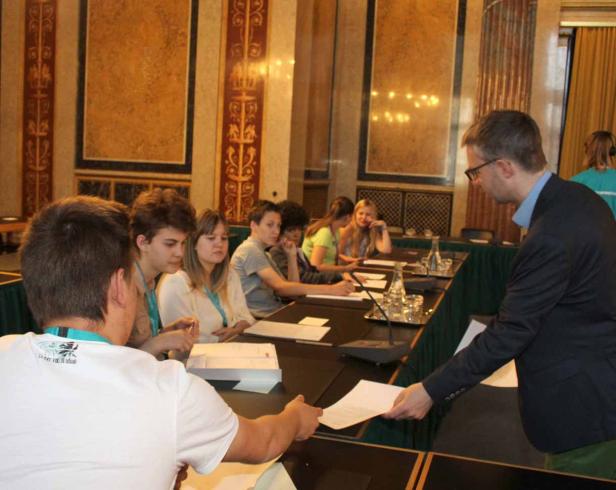 Ein Mann in Anzug übergibt ein Dokument an einen Jugendlichen in einem formellen Raum.