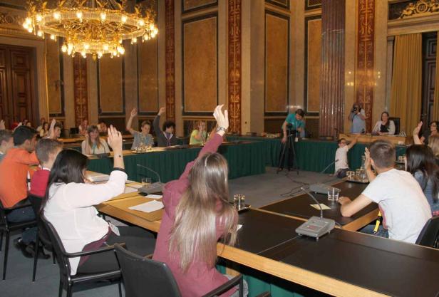 In einem prunkvollen Saal heben mehrere Personen ihre Hände.