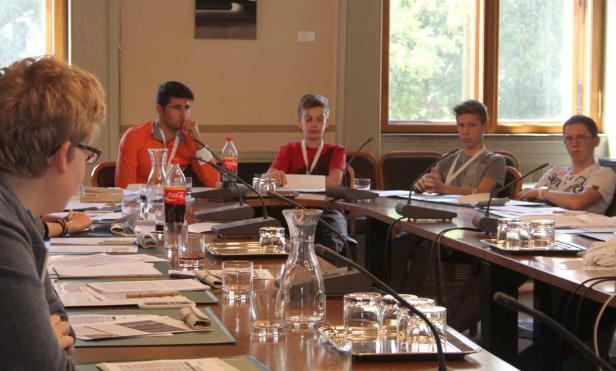 Mehrere Jugendliche sitzen an einem Tisch mit Mikrofonen und Gläsern bei einer Konferenz.