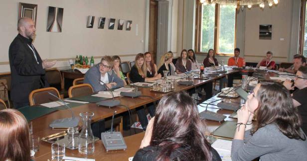 Ein Mann hält einen Vortrag vor einer Gruppe von Zuhörern an einem langen Tisch.