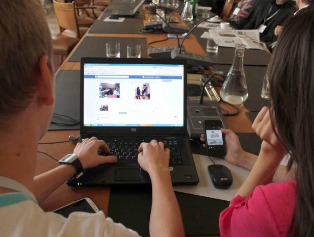 Zwei Personen sitzen an einem Tisch, eine tippt auf einem Laptop, die andere schaut auf ein Smartphone mit dem Logo „Demokratie Werkstatt“.