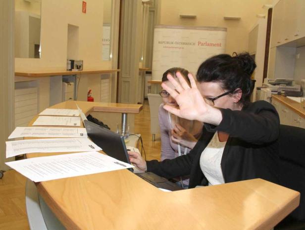 Eine Frau hält ihre Hand vor das Gesicht, während sie an einem Laptop arbeitet; im Hintergrund eine weitere Person und ein Banner des österreichischen Parlaments.