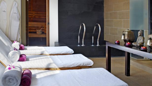 Innenansicht eines Spa-Bereichs mit Liegen, Sauna und einem kleinen Buddha auf einem Tisch.