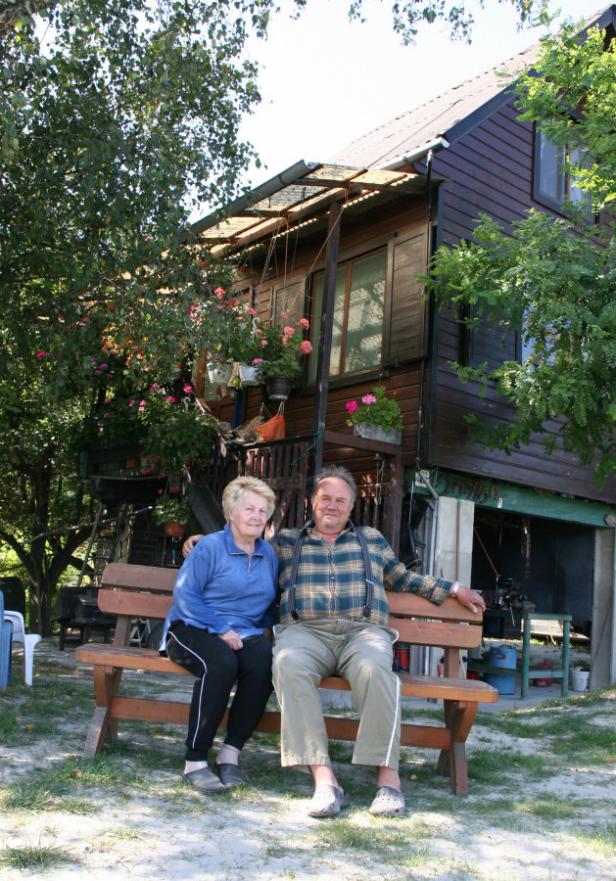 Daubelfischer und Angler Horst Steffek mit Gattin… Ein älteres Paar sitzt auf einer Bank vor einem hölzernen Haus mit Blumenkästen.