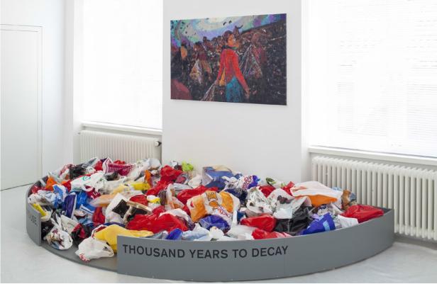 Eine Installation mit Plastiktüten unter einem Gemälde mit Menschen und dem Schriftzug „Thousand Years to Decay“.