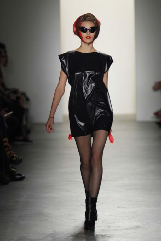 Eine Frau mit Sonnenbrille und Kopftuch trägt ein glänzendes, schwarzes Kleid und Stiefel auf dem Laufsteg.