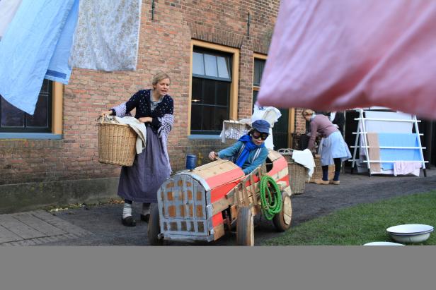 Frauen in traditioneller Kleidung verrichten Hausarbeiten, während ein Kind in einem Spielzeugauto sitzt.