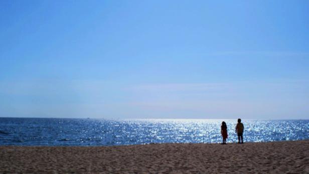 Zwei Personen stehen am Strand und blicken auf das glitzernde Meer.