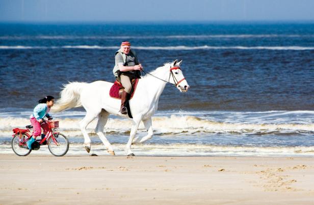 Ein Mann reitet auf einem weißen Pferd am Strand, während ein Mädchen auf einem Fahrrad nebenherfährt.