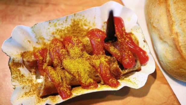 Eine Currywurst mit Ketchup und Currypulver in einer Pappschale, daneben ein Brötchen.