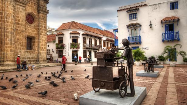 Eine belebte Plaza mit einer Skulptur, Tauben und traditioneller Architektur in Cartagena, Kolumbien.