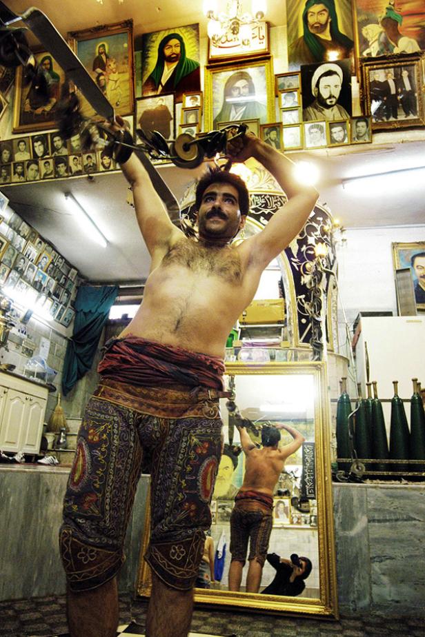 Ein Mann trainiert mit Keulen in einem traditionellen iranischen Fitnessstudio, geschmückt mit Porträts.