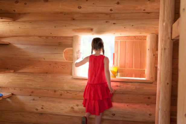 Ein Mädchen im roten Kleid schaut aus einem Fenster in einem Holzhaus.