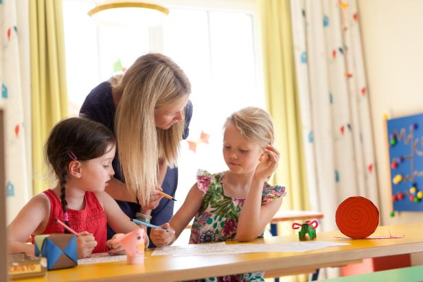 Eine Frau hilft zwei Mädchen beim Malen und Basteln an einem Tisch.