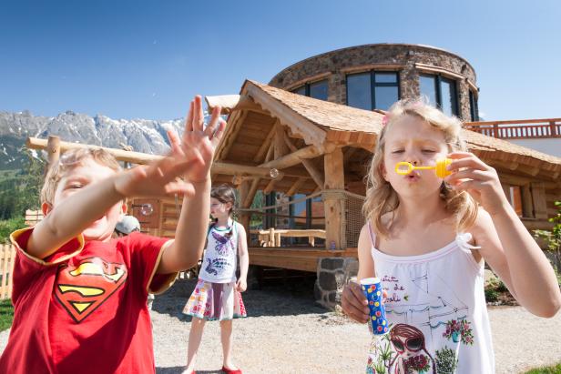 Kinder spielen im Freien und machen Seifenblasen vor einer Holzhütte.