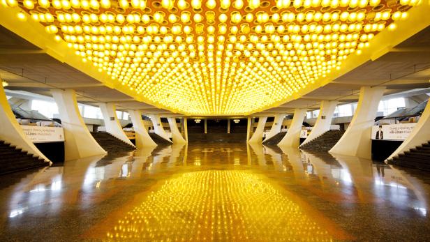 Ein großer Saal mit reflektierendem Boden und einer Decke voller Lampen.