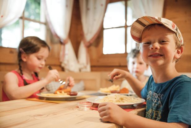 Drei Kinder sitzen an einem Holztisch und essen Nudeln und Pommes.