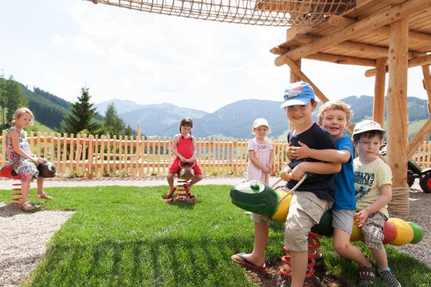 Kinder spielen auf einem Spielplatz mit Schaukeltieren vor einer Bergkulisse.